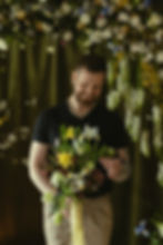 Man smiling, holding a bouquet of colorful flowers, standing against a backdrop of hanging blooms and green drapery, creating a joyful vibe. Photo by Lada Jensen