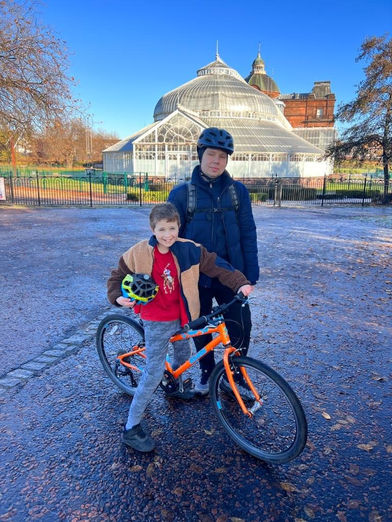 Buzz Cycling instructor stands together with a boy who just learned how to cycle. Both are smiling. They are standing in front of People's Palace in Glasgow Green.