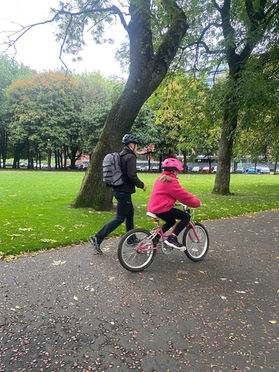 A girl in a pink jacket wears a pink helmet and cycles in the park. The instructor in a black helmet walks along her