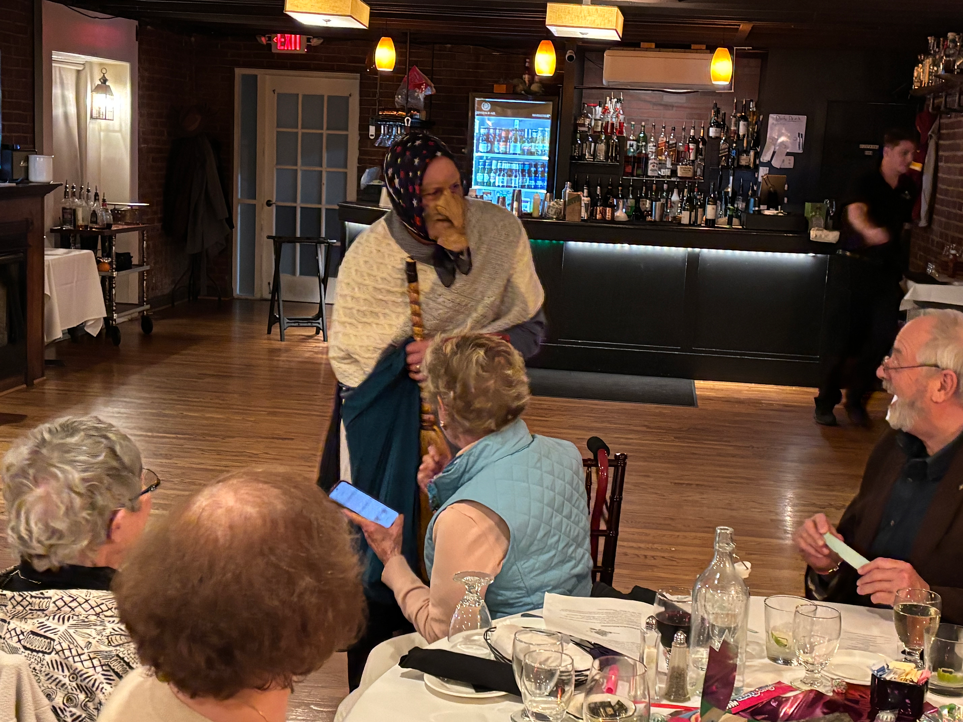 Costumed person with long nose, headscarf, broomstick entertains dinner guests.