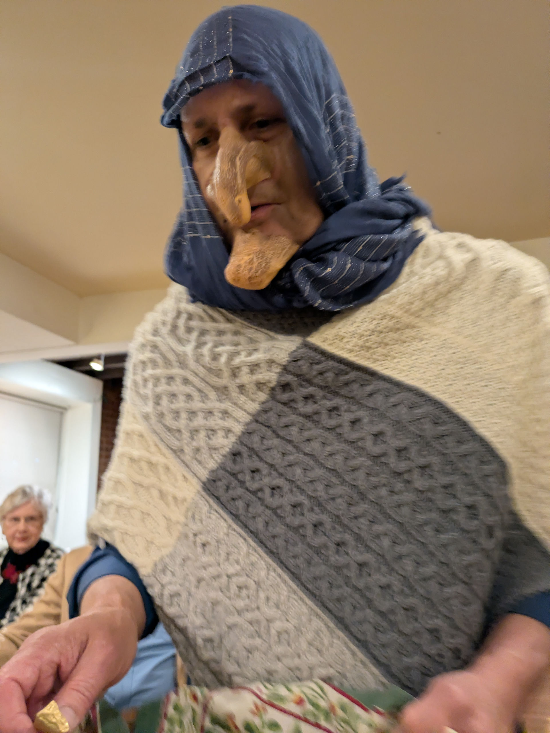 Character in costume with large prosthetic nose, blue headscarf, patterned poncho.