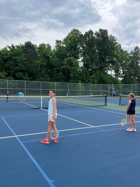 Girls playing tennis on a blue court with green trees background.