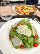 Arugula salad with cherry tomatoes, parmesan shavings, blurred pizza, and wine glass.