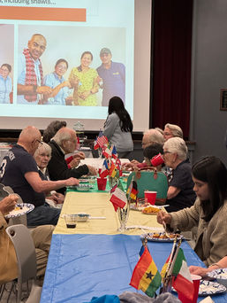 Diverse group enjoying potluck meal with international flags on table.