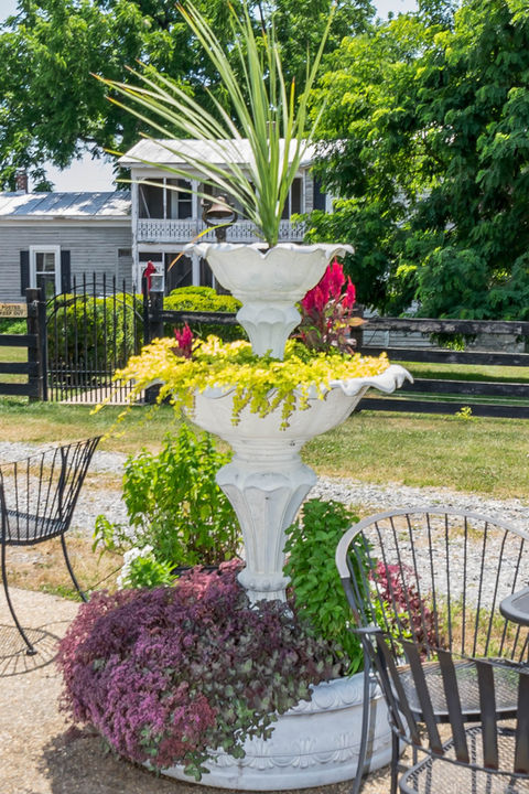 Two-tiered white planter with various colorful plants near a grey house.