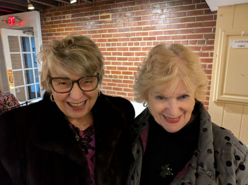 Two smiling women, one wearing glasses, against a brick wall.