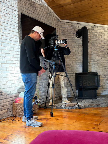 Two men filming indoors operating camera on tripod, brick fireplace background.