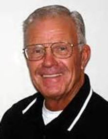 Portrait of Steve Souder wearing glasses and a black collared shirt with white trim, smiling in front of a plain white background.