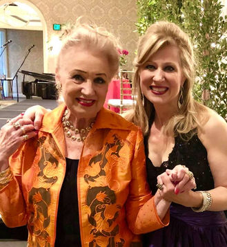 Two smiling women pose for photo, wearing elegant evening attire, holding hands.
