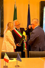 Bald man in Italian sash presents item to another man, woman watches.