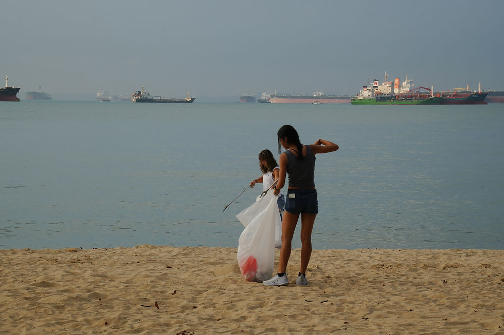 Two people picking up trash on a sandy beach with ships.