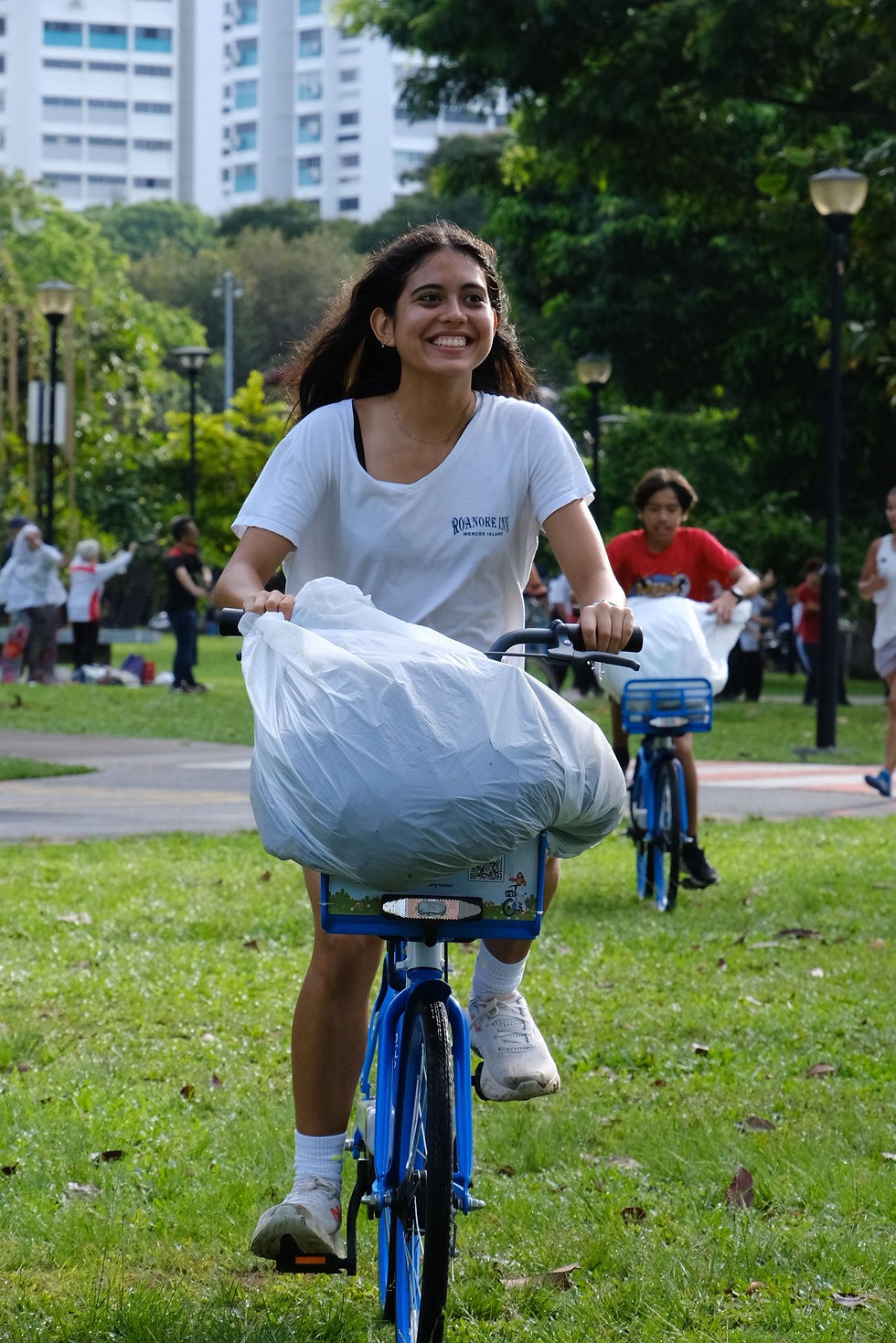 Smiling woman rides a blue bike with a large white bag in a park