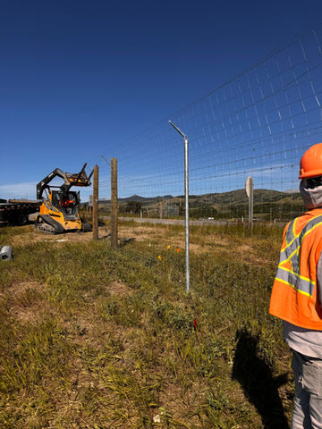 Worker observes the excavator setting fence posts on a sunny construction site.