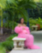 Pregnant woman in a pink maternity gown seated on a stone bench in a tropical garden.