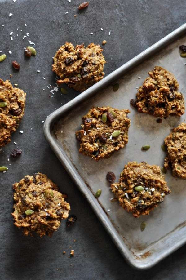 Baked cookies scattered, some on baking sheet and others on dark surface with pumpkin seeds and dried fruit sprinkled around it