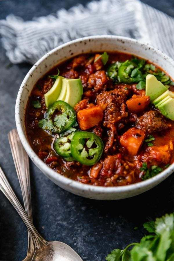 A close up shot of a white bowl on a dark surface that's filled with Slow Cooker Chunky Beef Chili with Sweet Potatoes and topped with sliced jalapeños, avocado, and fresh cilantro.