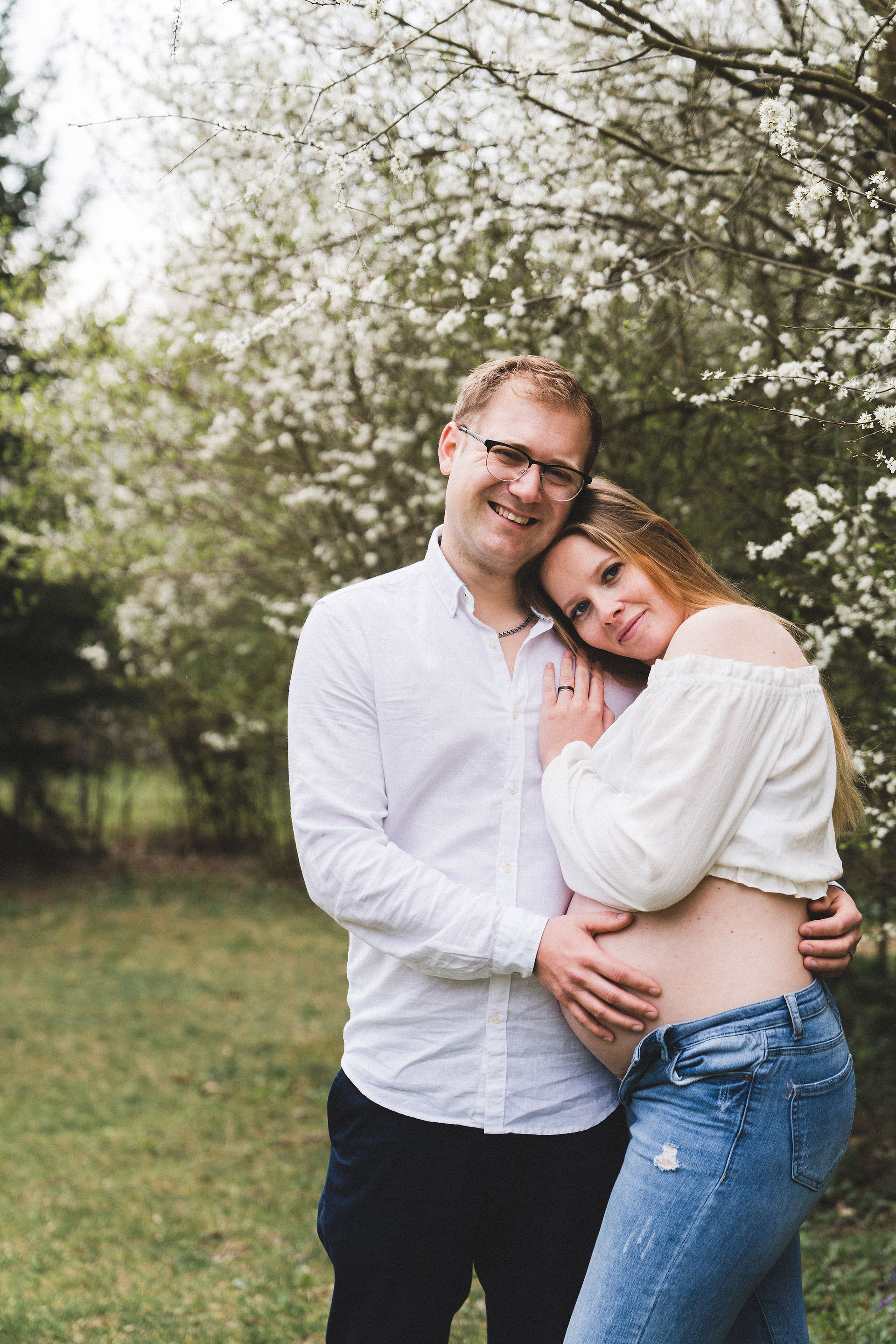 Werdende Mama und Papa in natürlicher Pose, beide Hände auf dem Babybauch, stehen zusammen im Freien.