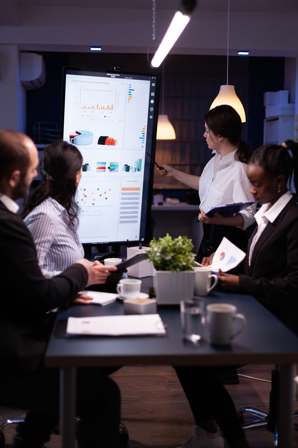 People in formal attire discuss data charts on a screen in an office. A woman points at graphs. Modern, professional atmosphere.