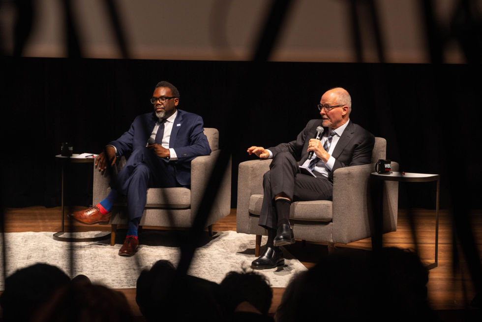 Two men in suits, Brandon Johnson and Paul Vallas seated on a stage in a discussion debate at a public event at University of Chicago. The audience is in the foreground silhouette. Photo by Matt Petres