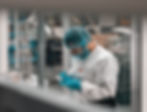 Person in lab gear examines samples in lab setting. Wearing blue gloves, hairnet, and white suit. Focused demeanor; "3M" visible.