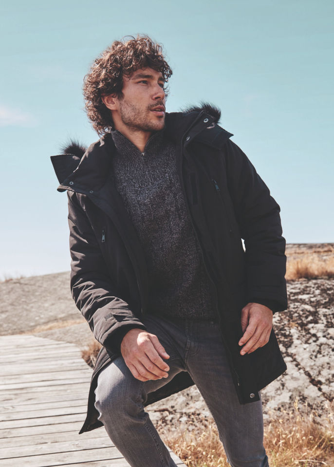 Gémo "Winter" seasonal campaign shot in Sweden featuring a man in a black technical parka and grey knit sweater on a coastal boardwalk. French fashion brand outdoor lifestyle art direction and cold-weather menswear styling.