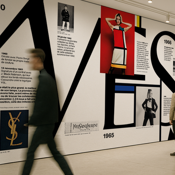 Yves Saint Laurent exhibition mural featuring a large-scale typographic timeline with historical fashion photography and iconic logo designs. Luxury fashion scenography and historical art direction for a gallery installation.