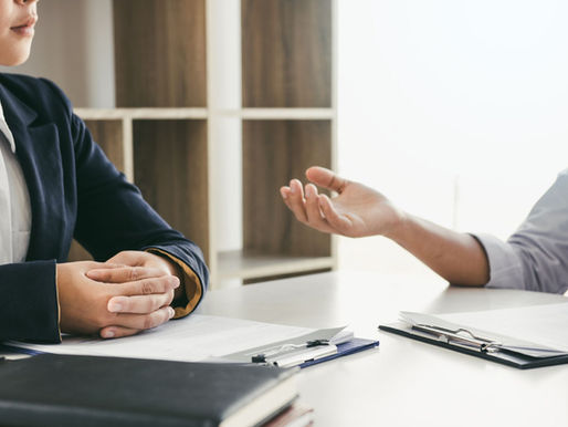 Two professionals in business attire seated at a table during a serious workplace conversation, one gesturing while speaking and the other listening with hands clasped — illustrating a manager giving an employee feedback about a promotion decision.