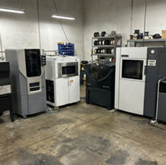 Impressora 3D industrial Stratasys parada em galpão com cobertura de proteção.