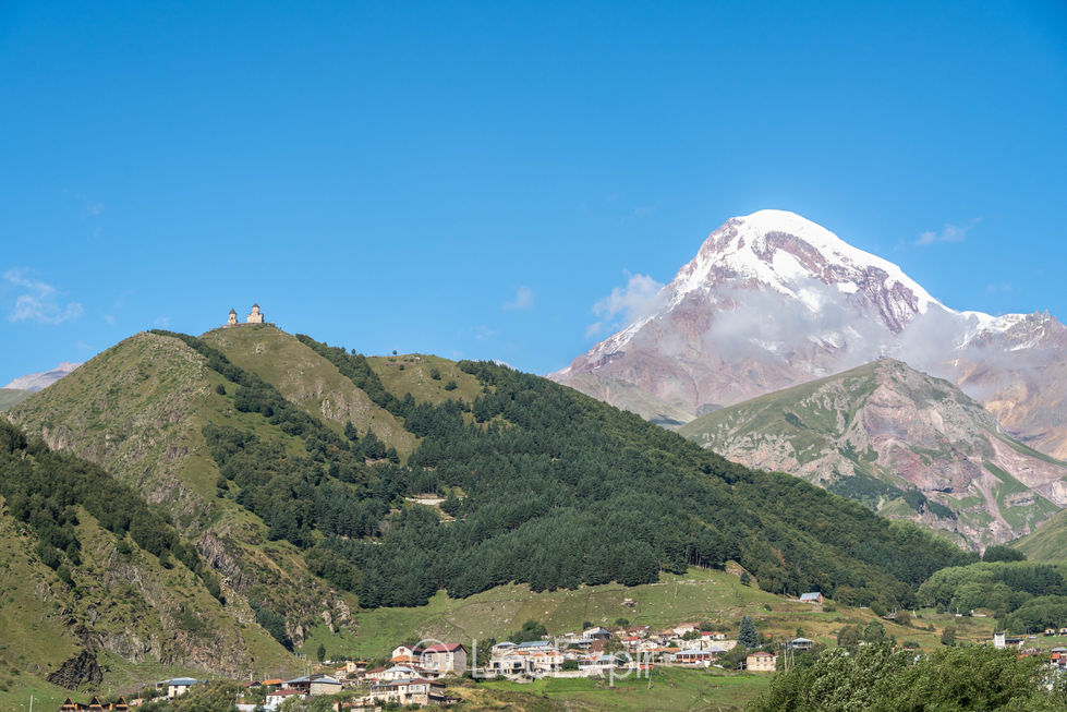 Snow-capped mountain, Mount Kazbek, rises behind lush green hills in the Caucasus with the Gergeti Trinity Church on top and the small village of Gergeti below, beneath a clear blue sky, creating a peaceful alpine landscape. Kazbegi in Georgia.