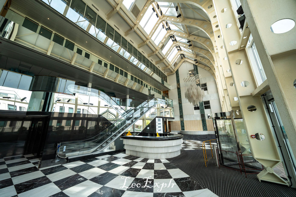 Spacious atrium with high arched ceilings, glass walls, and checkered floor. Central round desk, escalators, and grand chandelier create an elegant ambiance.