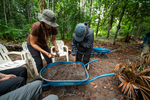 Two people in outdoor clothing examine soil in a blue wheelbarrow amidst a lush forest. Empty chairs surround them, suggesting a group activity. In the Amazon Rainforest in Brazil.