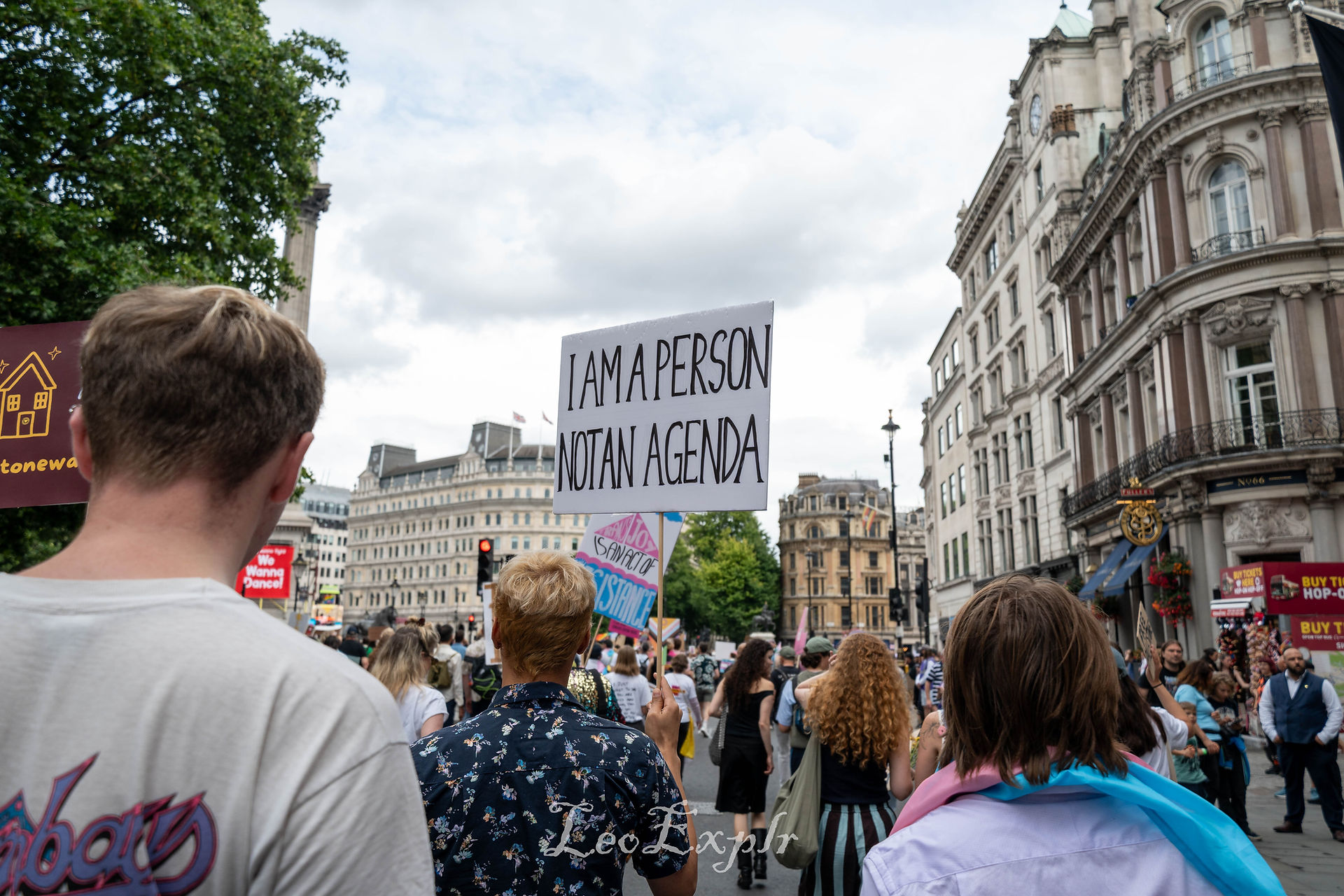 London Trans Pride. A crowd of people marches down a city street. A person holds a sign reading "I am a person, not an agenda," conveying a tone of protest and advocacy.