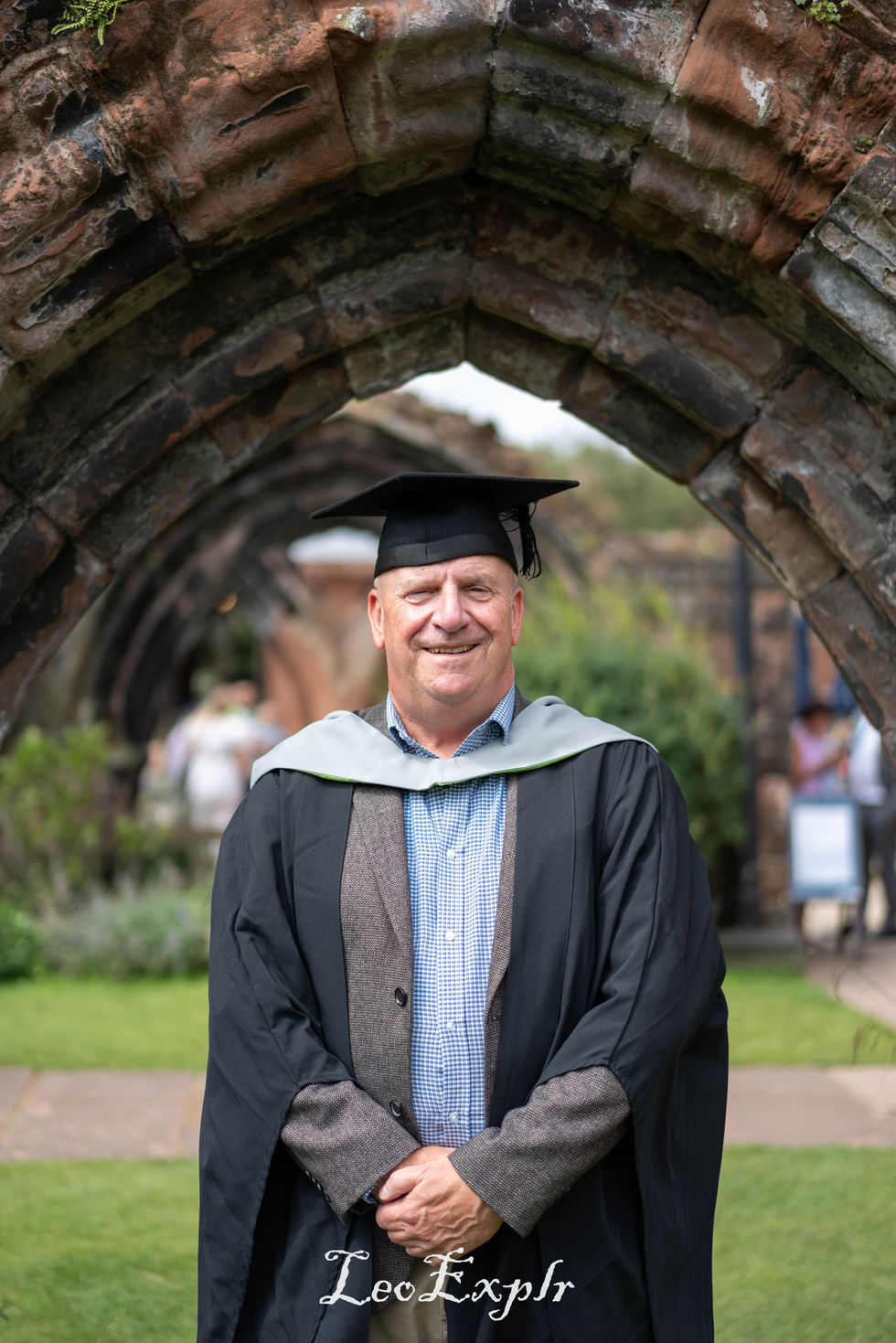 A man in a graduation gown and cap stands smiling under a stone archway. His expression is proud and joyful, with a sunny garden in the background.