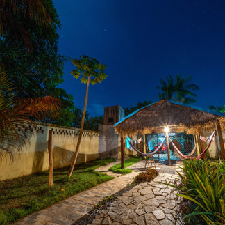 Nighttime garden scene with a lit stone pathway leading to a thatched-roof hut. Hammocks hang inside, surrounded by lush greenery and a starry sky. IPBio volunteer house in Caranazal community near Alter do Chao in Brazil