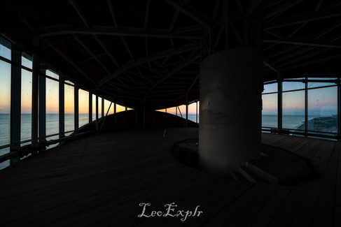 Anaklia abandoned tower, Georgia. Dimly lit interior of an empty structure with large windows revealing a serene ocean view at sunset. A central pillar and exposed ceiling beams add to the minimalist atmosphere.