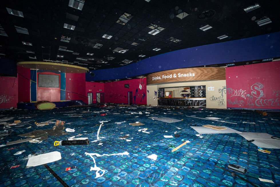 Abandoned, graffiti-covered entertainment hall lobby with scattered debris. A closed "Drinks, Food & Snacks" counter sits amid a dark, eerie atmosphere. Chatham Buzz Bingo.