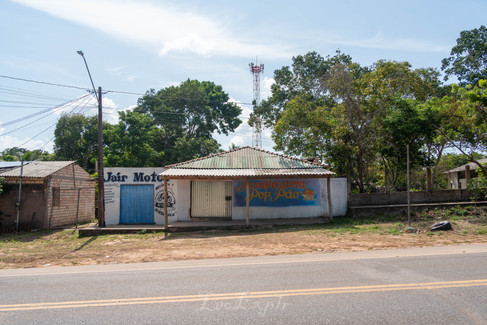 Small roadside store with paint-covered walls, blue and white doors, surrounded by lush trees under a clear sky. A utility pole and telecom tower are visible. Bakery in the Caranazal community near Alter do Chao, Brazil.