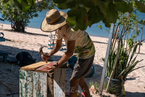 Man in a straw hat working on a wooden project by the beach, surrounded by sand and plants. Sunlit scene conveys a calm, tropical vibe. Ilha do Amor in Alter do Chao, Brazil.
