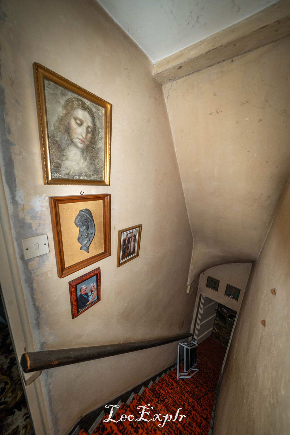 Narrow stairwell with red patterned carpet and beige walls displaying framed art, including a portrait inside an abandoned house. Cosy, vintage atmosphere.