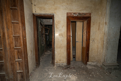 Sanatorium Meshakhte in Tskaltubo, Georgia. Abandoned interior with two wooden doorways, peeling paint, and debris on the floor. The dim lighting and neglected condition evoke a sense of decay.