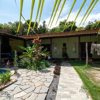 A serene backyard scene features a pool, lush tropical plants, and a small green-roofed house under a blue sky. Laundry hangs and sunlight filters through. IPBio volunteer house in Caranazal community near Alter do Chao in Brazil