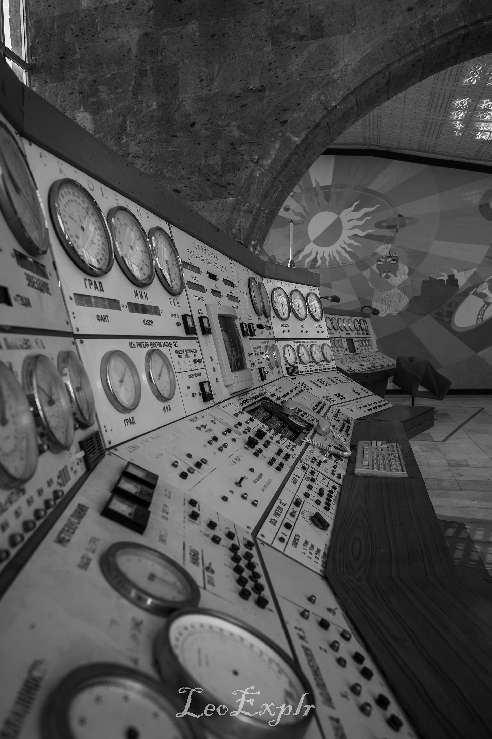 Aragats Scientific Center - Orgov Radio-Optical Telescope. Black and white photo of an old control room. A row of dials and switches lines the wall beneath an arch. A colourful mural is partially visible.
