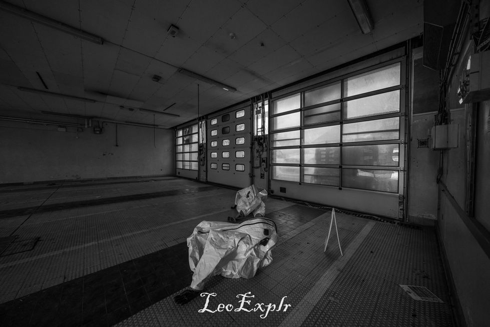 Black and white image of an abandoned garage with two worn hazmat suits lying on the floor. Sunlight filters through large, grimy windows, creating an eerie and desolate atmosphere.