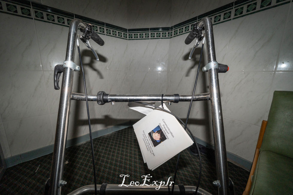 A metal walker stands in a dimly lit corner of a tiled room. A paper 'Communications Passport' for 'David Walker' with text and an image hangs from the handlebar, giving a somber, neglected feel. Beech Lodge, Horsham.
