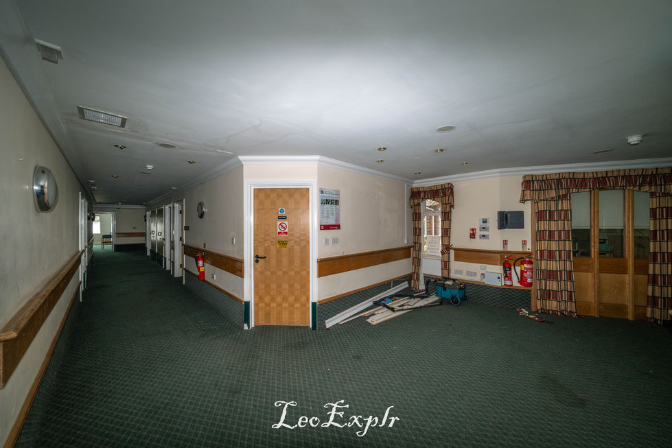 Dimly lit, empty nursing home hallway with patterned carpet, two wood-paneled doors, fire extinguisher, and scattered equipment creating an abandoned feel. Beech Lodge, Horsham.