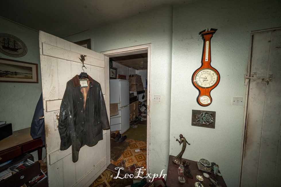 An old coat hangs on a worn, white wooden door in a cluttered room. A vintage barometer is mounted on a pale wall, evoking a nostalgic, abandoned atmosphere.