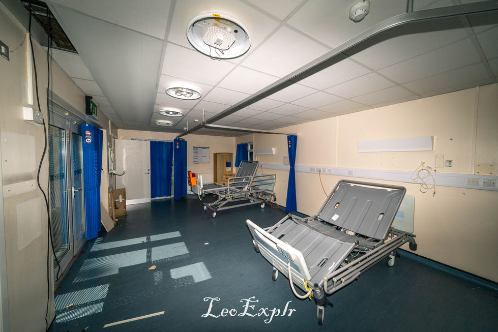 Empty hospital room with two adjustable beds, blue curtains, and overhead lights. The space feels abandoned and stark with minimal lighting. Edenbridge and District War Memorial Hospital.