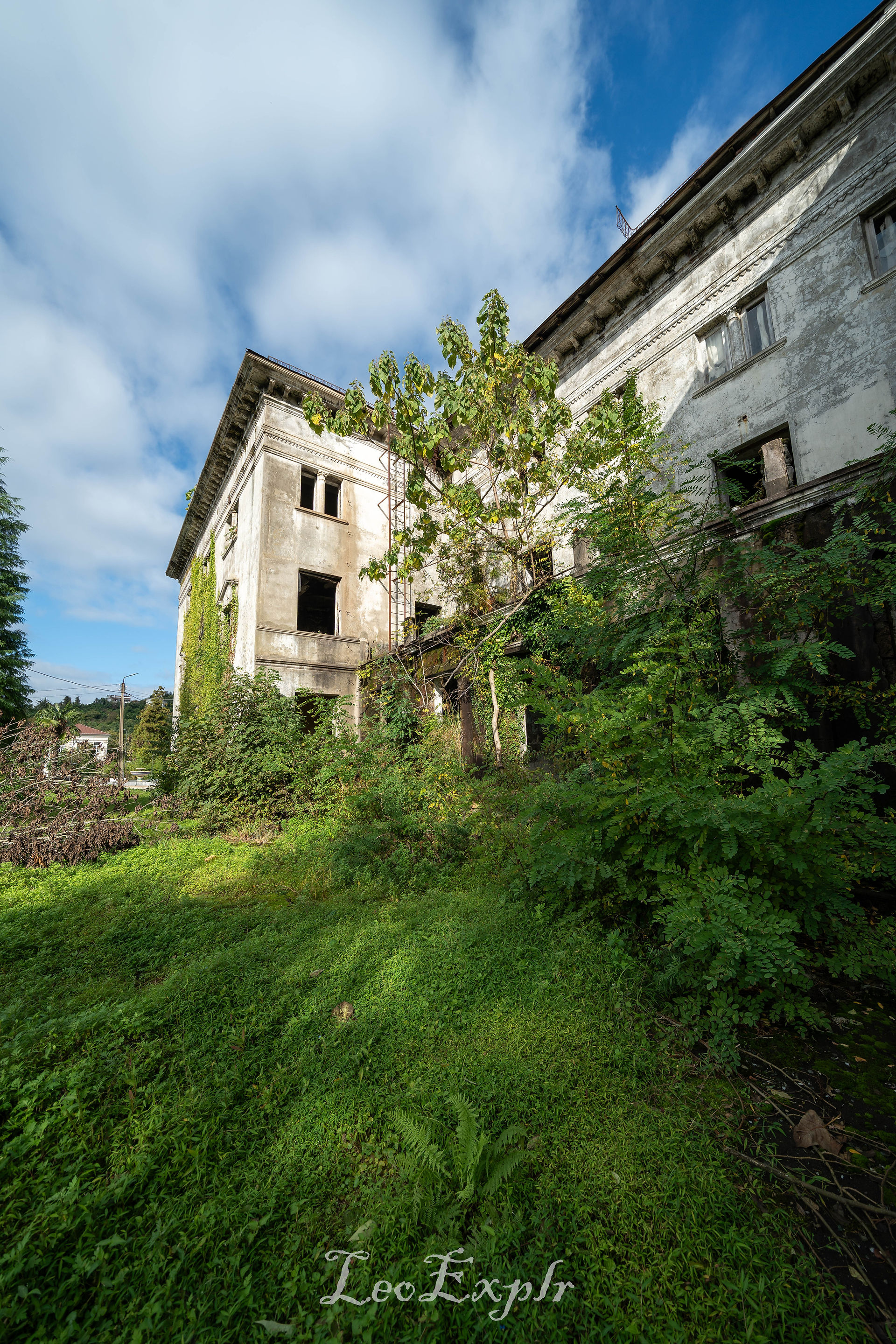 An abandoned, overgrown building with cracked walls and broken windows, surrounded by lush green plants under a blue sky with scattered clouds. Former Palace of Culture in Dagva, Georgia.