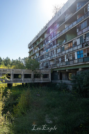 Hotel Aia in Tskaltubo, Georgia. A large, abandoned building with numerous broken windows and weathered, colorful panels. It's surrounded by overgrown greenery under a clear blue sky.