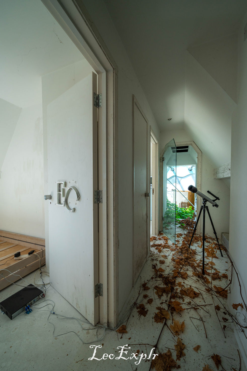 A narrow, cluttered hallway with white walls is scattered with dry leaves. A telescope on a tripod stands near an open door, adding an eerie, abandoned feel. Butterfly House in Surrey.